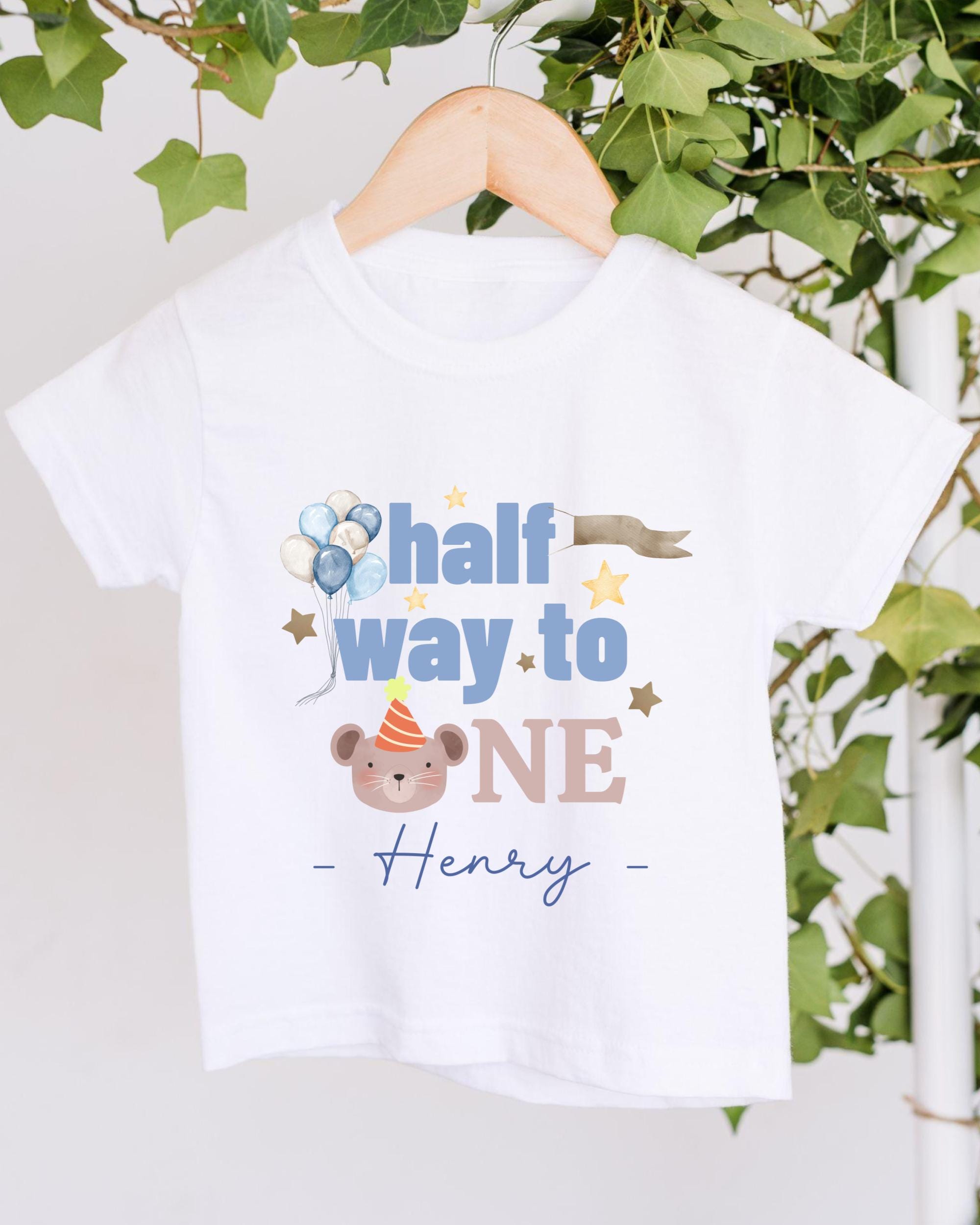 A white t-shirt with a graphic design featuring a cartoon bear wearing a party hat and balloons, along with the text "half way to one Henry" printed on it.
