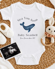 a baby announcement, including a white onesie with a blue whale graphic, a crocheted bunny toy, and a baby announcement photo.