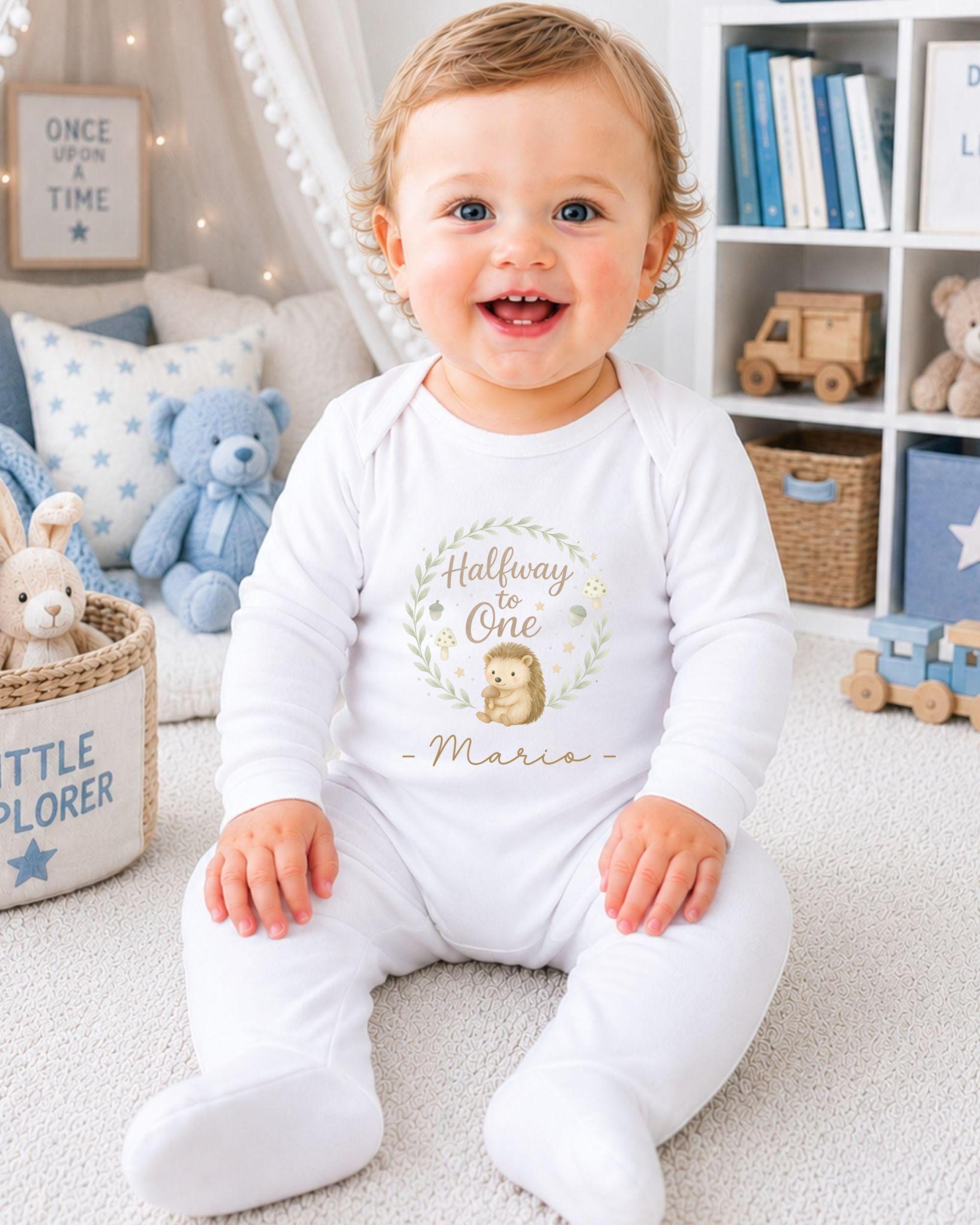 A smiling baby boy wearing a white onesie with a hedgehog design and the text "Halfway to one" is sitting on the floor in a nursery setting.