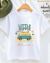 A white t-shirt with a colorful graphic design featuring a van, palm trees, mountains, and a sun. The text on the t-shirt says "Little Hippie Oliver".