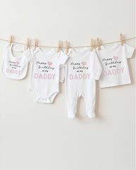 A collection of four white baby clothes, including onesies and bibs, hanging on a clothesline. Each piece of clothing has a message printed on it, likely related to a birthday celebration.