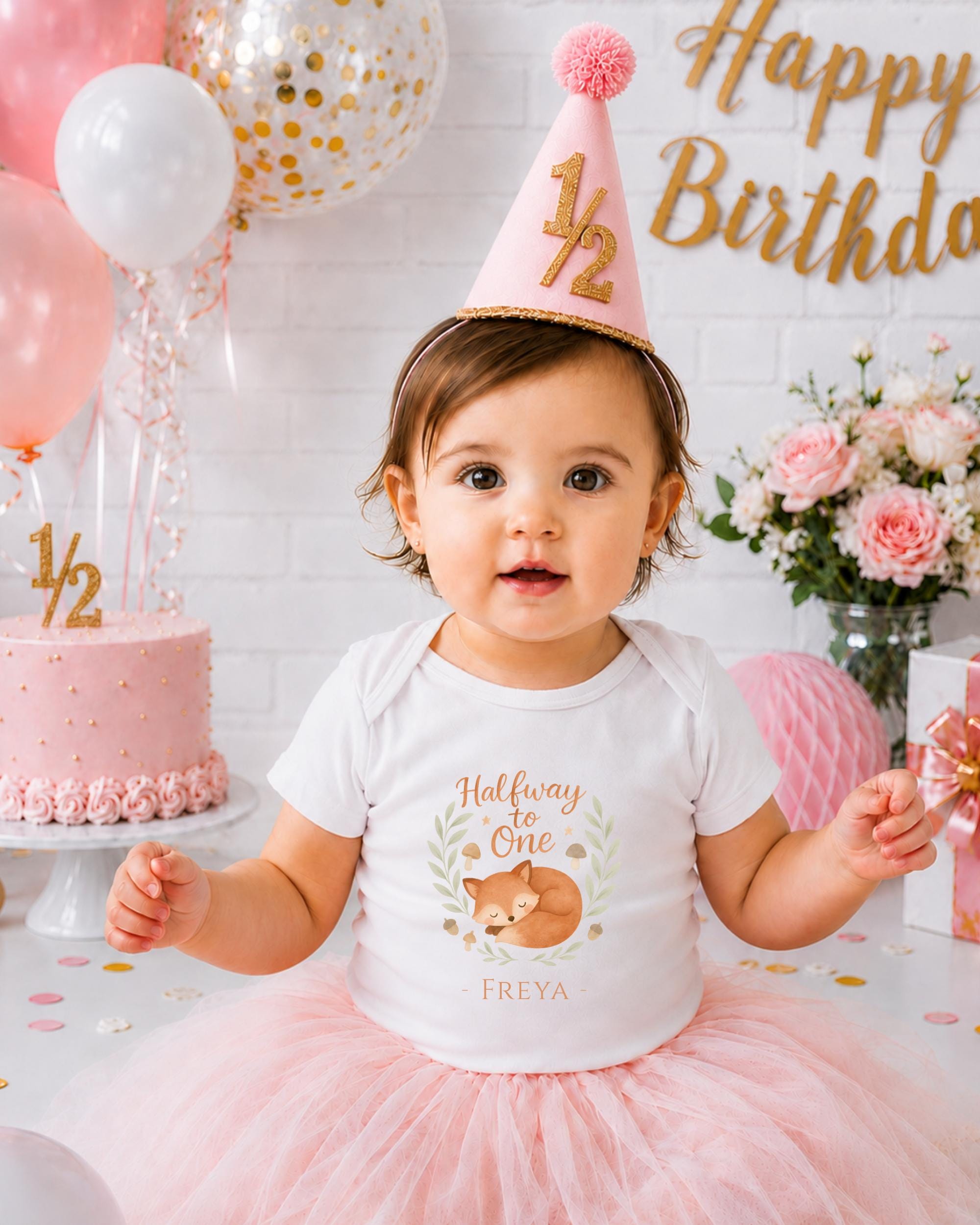 A young girl wearing a white t-shirt with a fox design and a pink party hat sits in front of a birthday cake and balloons, celebrating her first birthday.