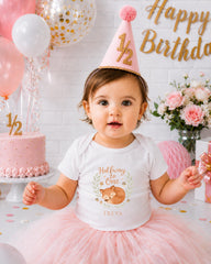 A young girl wearing a white t-shirt with a fox design and a pink party hat sits in front of a birthday cake and balloons, celebrating her first birthday.