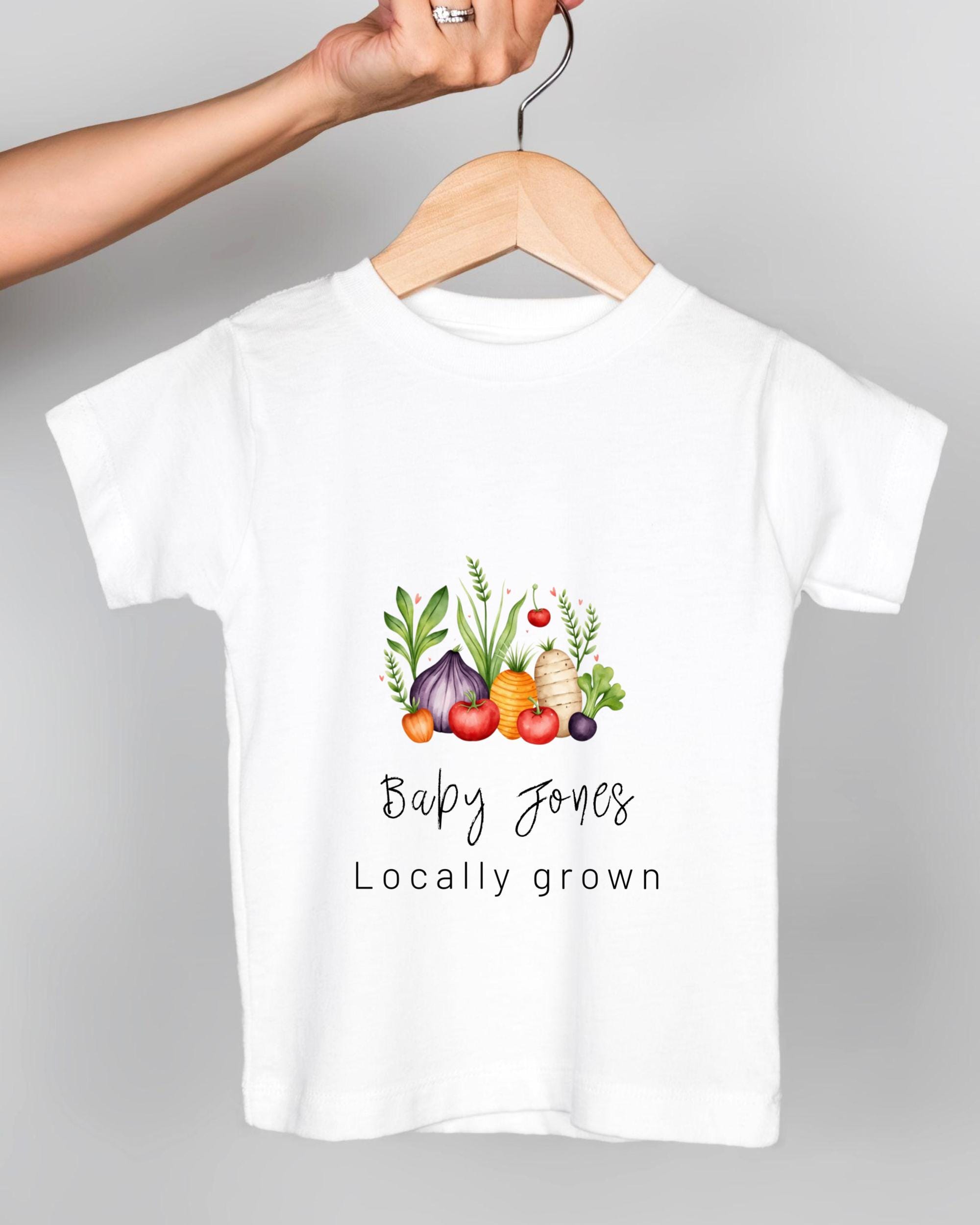 A white t-shirt with a graphic design of various vegetables hanging on a wooden hanger.