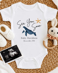 a baby announcement, including a white onesie with a blue whale and starfish design, a crocheted bunny toy, and a baby's firstborn announcement.