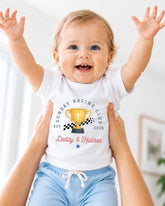 A smiling baby wearing a white t-shirt with a graphic of a trophy and the text "Sunday Racing Club Est. 2628" is being held up by two people.