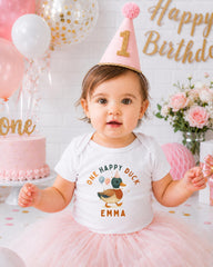 A young girl wearing a birthday hat and a white t-shirt with a duck design is posing in front of a birthday cake and balloons.