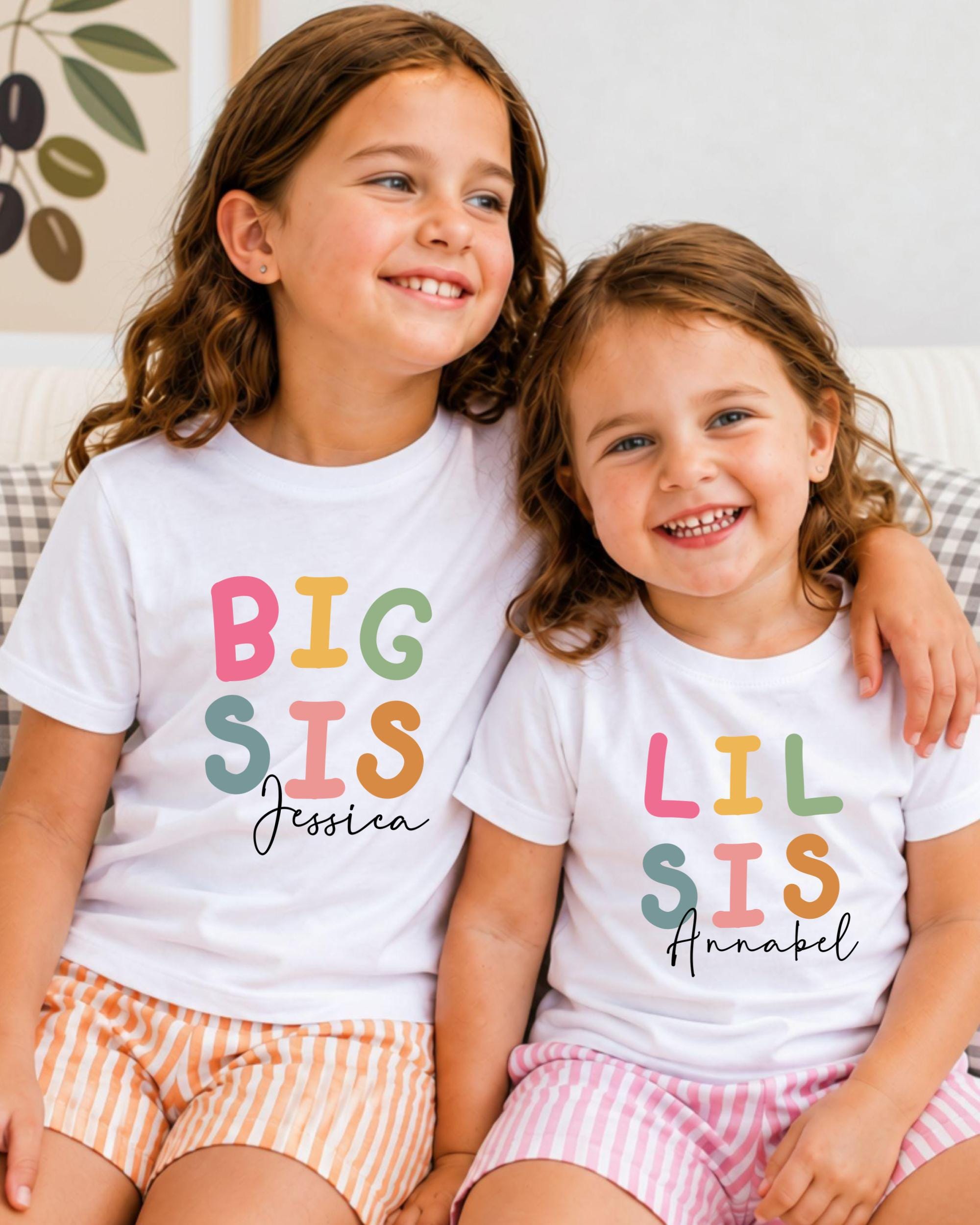 Two young girls wearing white t-shirts with colorful text and names on them, smiling and sitting together.