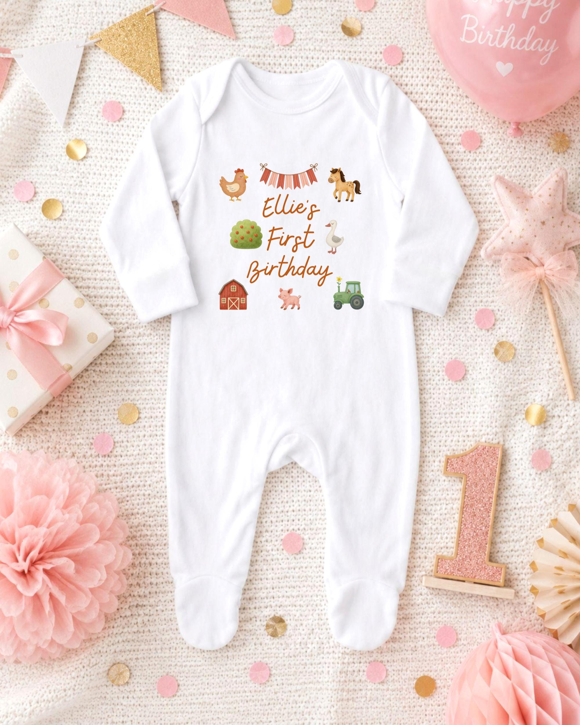 A white onesie with farm animal illustrations and the text "Ellie's First Birthday" is displayed on a bed with pink and gold confetti, a pink star, and a pink ribbon.
