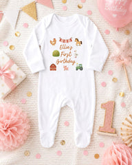 A white onesie with farm animal illustrations and the text "Ellie's First Birthday" is displayed on a bed with pink and gold confetti, a pink star, and a pink ribbon.
