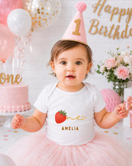 A young girl wearing a white t-shirt with a strawberry design and a pink birthday hat sits in front of a pink and white birthday cake, surrounded by balloons and flowers.