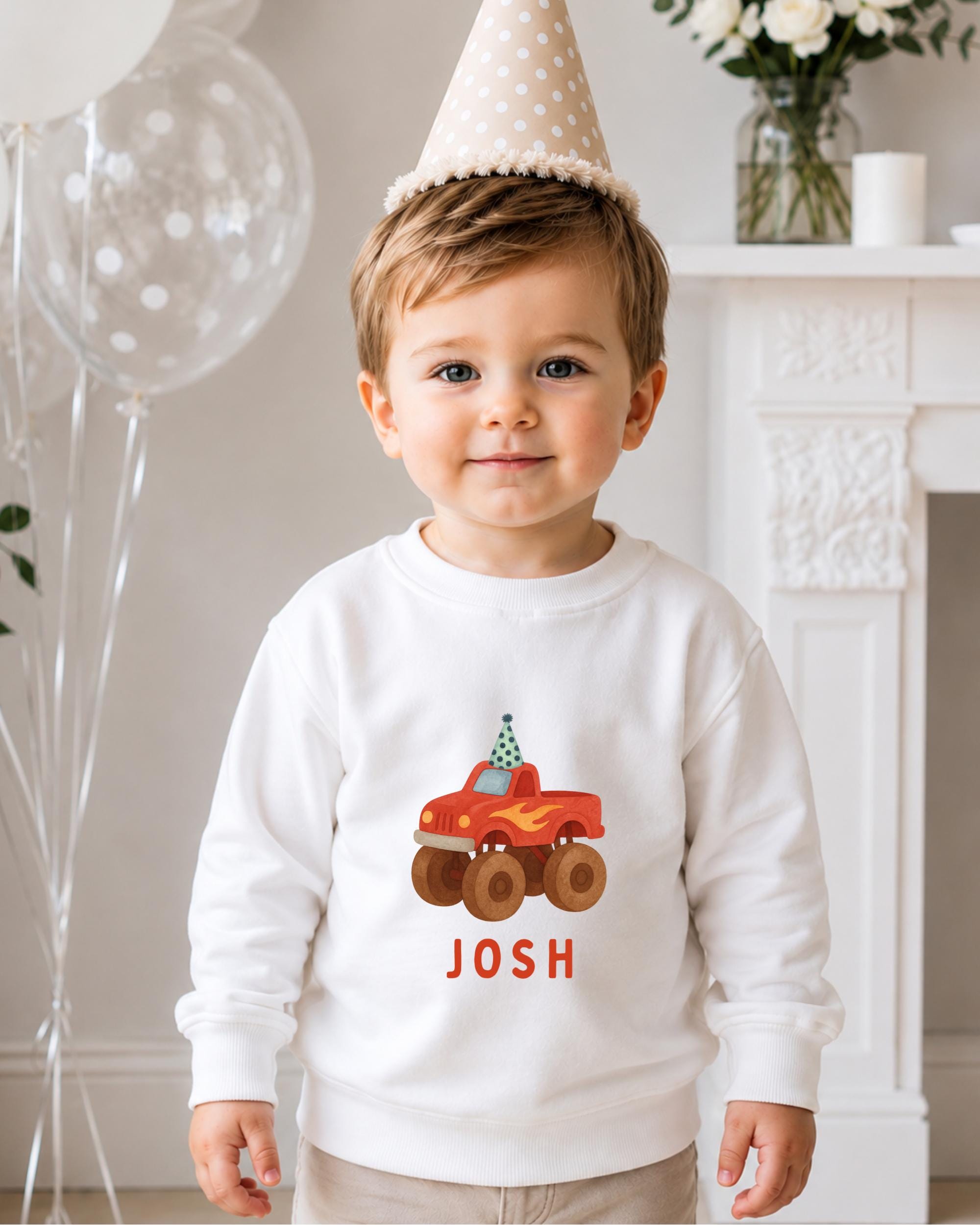 A young boy wearing a white sweater with a red truck and the name " Josh" printed on it, standing in front of a fireplace with balloons in the background.