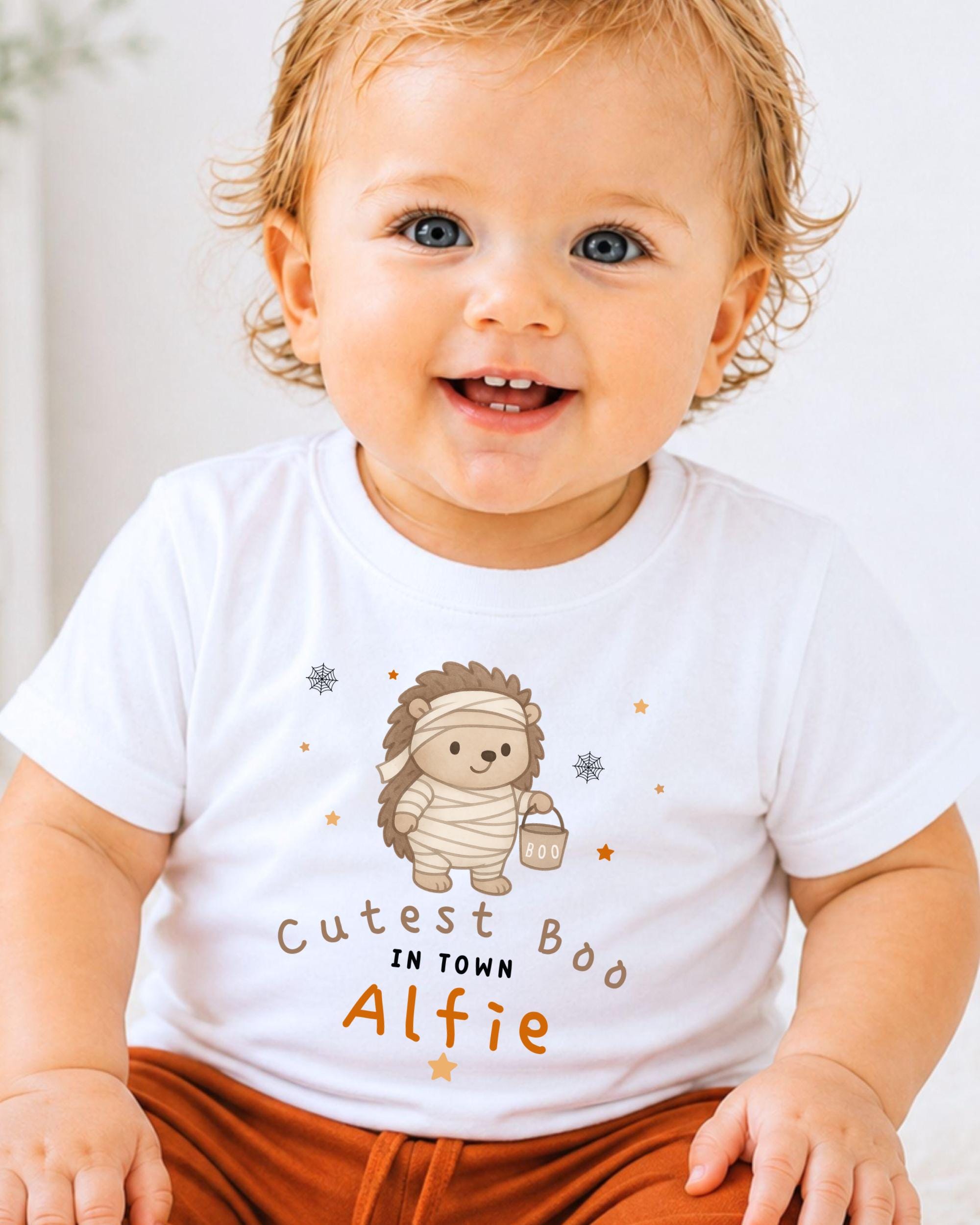 A smiling baby wearing a white t-shirt with a cartoon hedgehog character and text that says "Cutest Boo in Town Alfie".