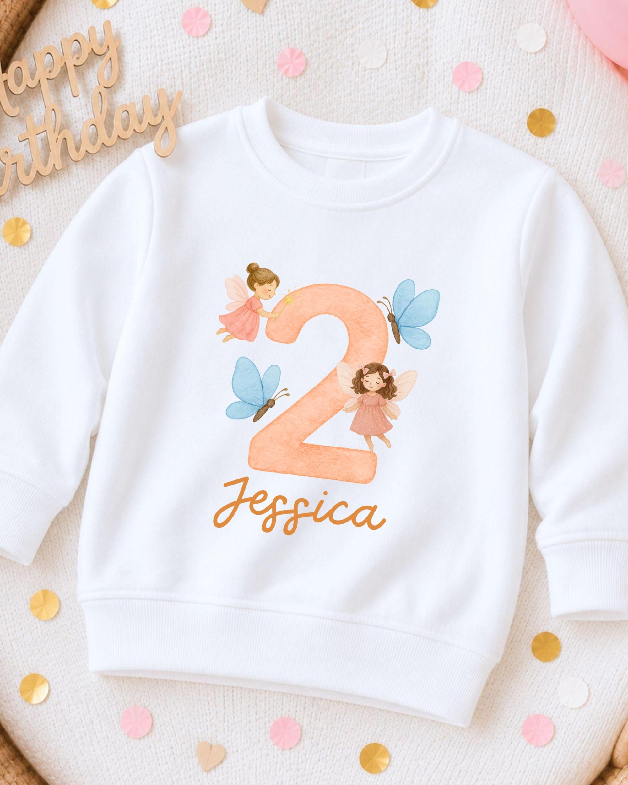 A white sweatshirt with a personalized number 2 and fairy-themed design, along with confetti and a "Happy Birthday" sign.