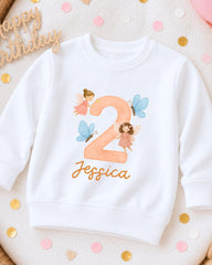 A white sweatshirt with a personalized number 2 and fairy-themed design, along with confetti and a "Happy Birthday" sign.