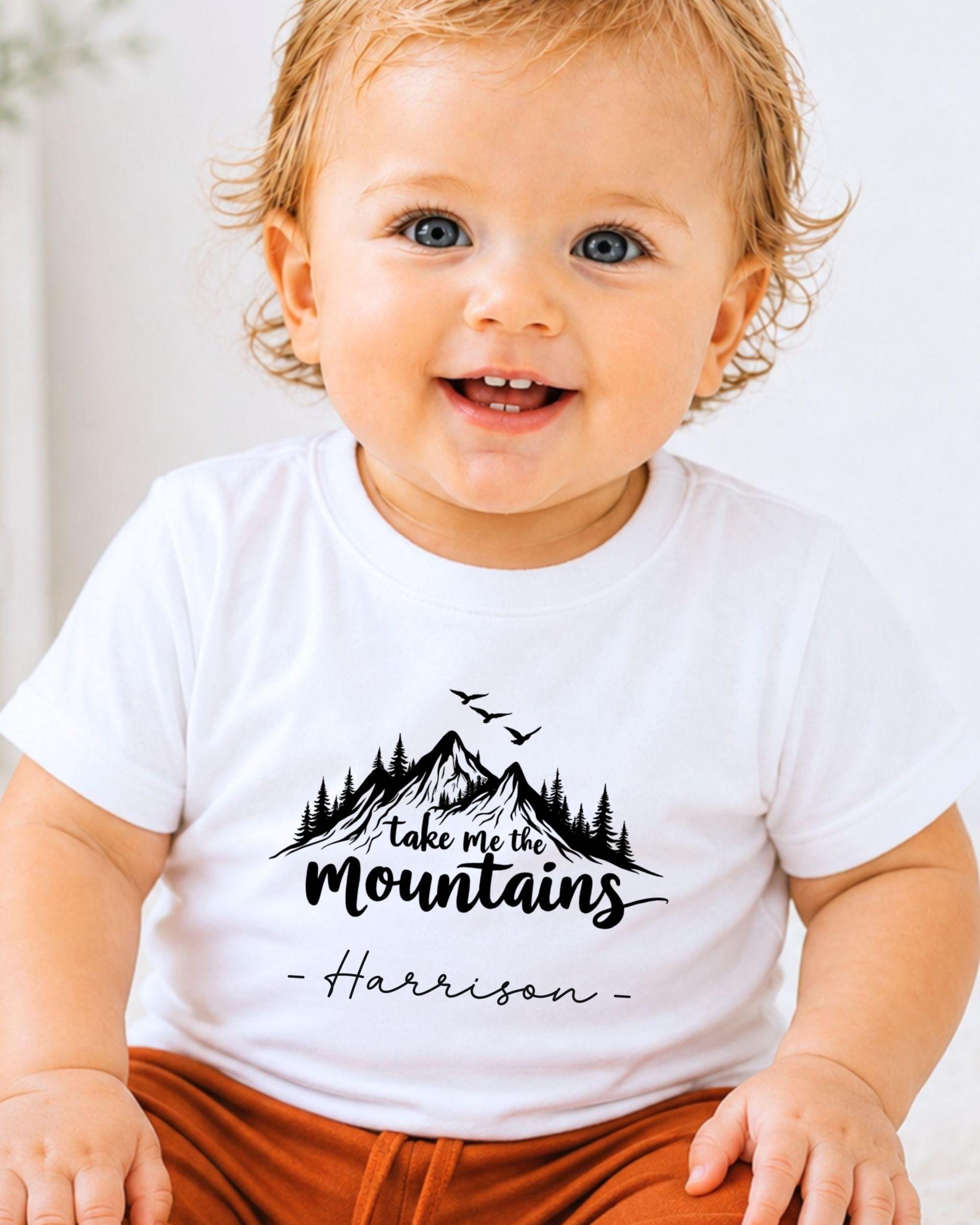 A smiling baby wearing a white t-shirt with a mountain graphic and the text "Take me the mountains" printed on it.