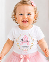 A smiling baby girl wearing a white t-shirt with a pink floral design and a pink bow in her hair.