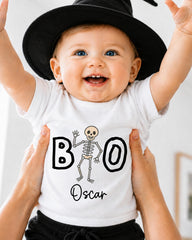 A smiling baby wearing a white t-shirt with a skeleton design and the name "Oscar" printed on it, being held up by someone's hands.