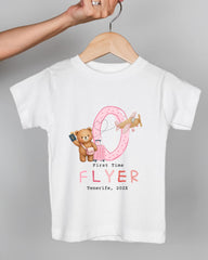 A white t-shirt with a graphic design featuring a teddy bear, a pink airplane, and the text "First Time FLYER Tenerife, 20XX" printed on it.