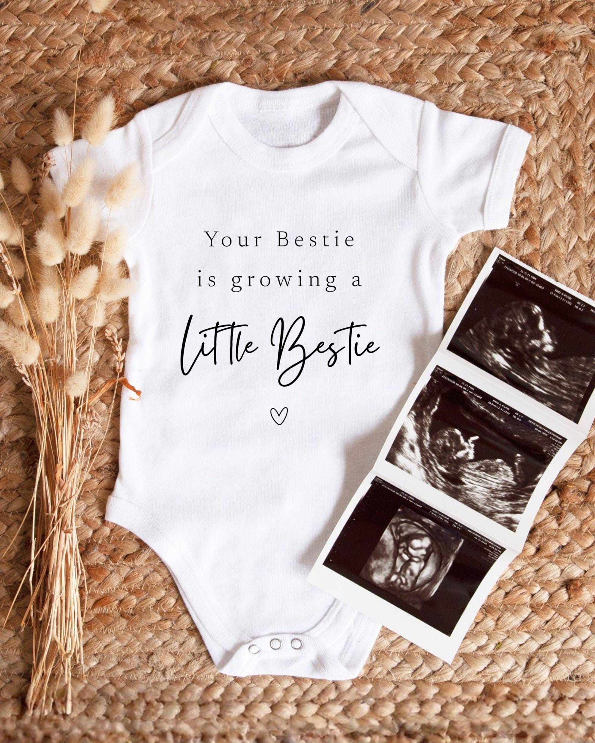 A white baby onesie with the text "Your Bestie is growing a Little Bestie" printed on it, along with a heart symbol. Next to the onesie is a set of three black and white images depicting an ultrasound image of a baby.