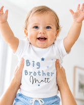 A smiling baby wearing a white shirt with the text "Big Brother Matt" and blue shorts, being held up by someone's arms.