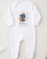 A white onesie with a graphic design on it, featuring a teddy bear, a passport, and an airplane. The text on the onesie says "First Time Flyer Hattie Barcelona, 202x".