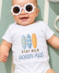 A smiling baby wearing a white onesie with a surfboard design and sunglasses, lying on a green and white striped surface.