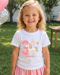 A young girl wearing a white t-shirt with a unicorn and balloons, smiling at the camera.