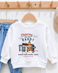 A white sweatshirt with a graphic design on it, hanging on a wooden hanger. The design features a cartoon image of a person cheering and a building with a clock tower. The text on the sweatshirt reads "Cheering for my DADDY" and "Marathon Name Here Custom Date".