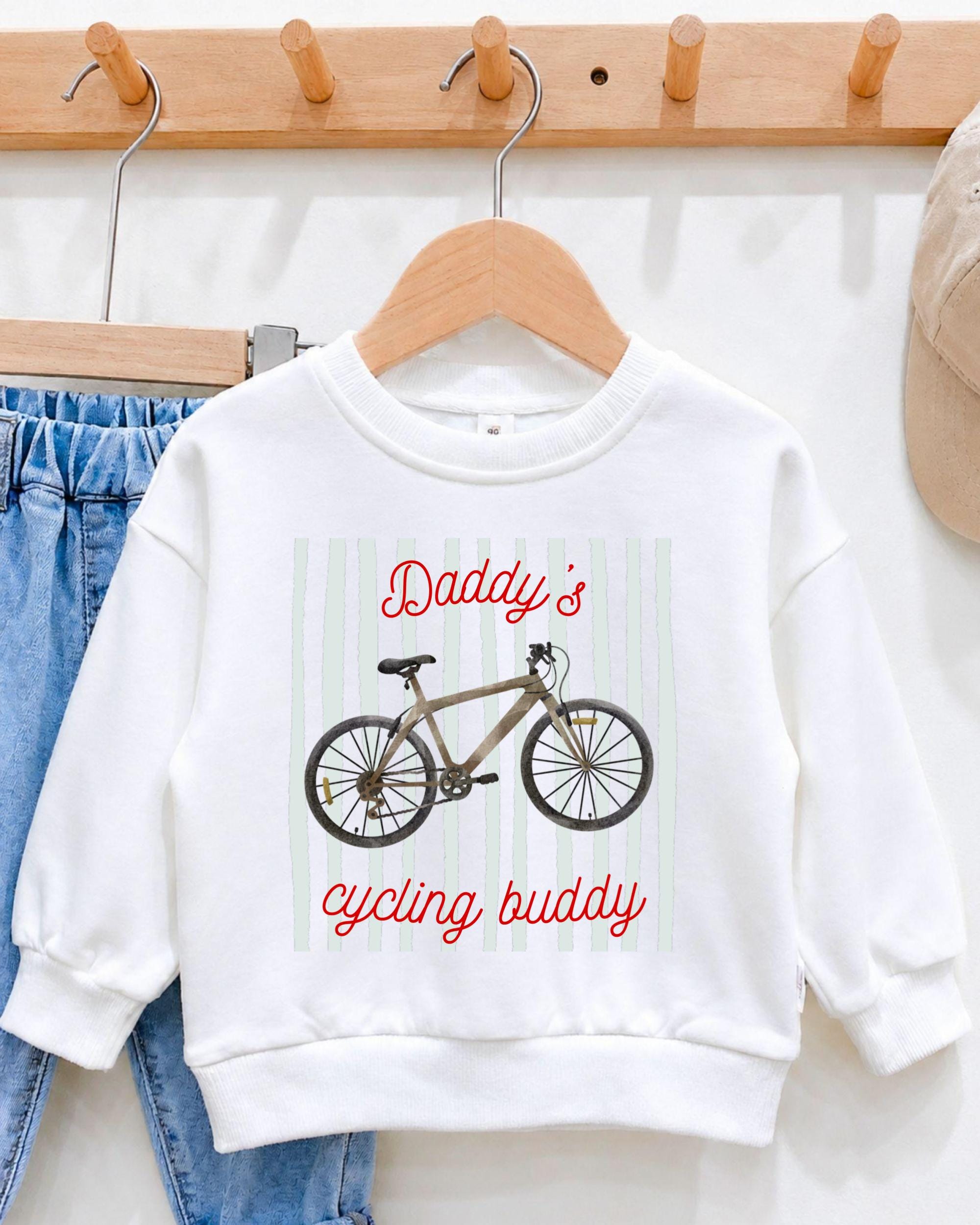 A white sweatshirt with a graphic of a bicycle and the text "Daddy's cycling buddy" printed on it, hanging on a wooden hanger.
