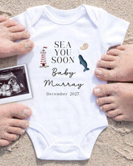 a white onesie with text and illustrations related to a baby announcement, surrounded by sand and a photograph of an Ultrasound image.