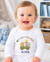 A smiling baby boy wearing a white onesie with a cartoon image of a car and animals, sitting in a room with a bookshelf and stuffed animals in the background.