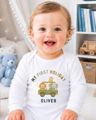 A smiling baby boy wearing a white onesie with a cartoon image of a car and animals, sitting in a room with a bookshelf and stuffed animals in the background.