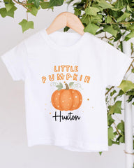 A white t-shirt with a pumpkin graphic and text printed on it, hanging on a wooden hanger.