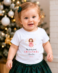 A smiling baby girl wearing a white t-shirt with a fairy graphic and text, standing in front of a Christmas tree.