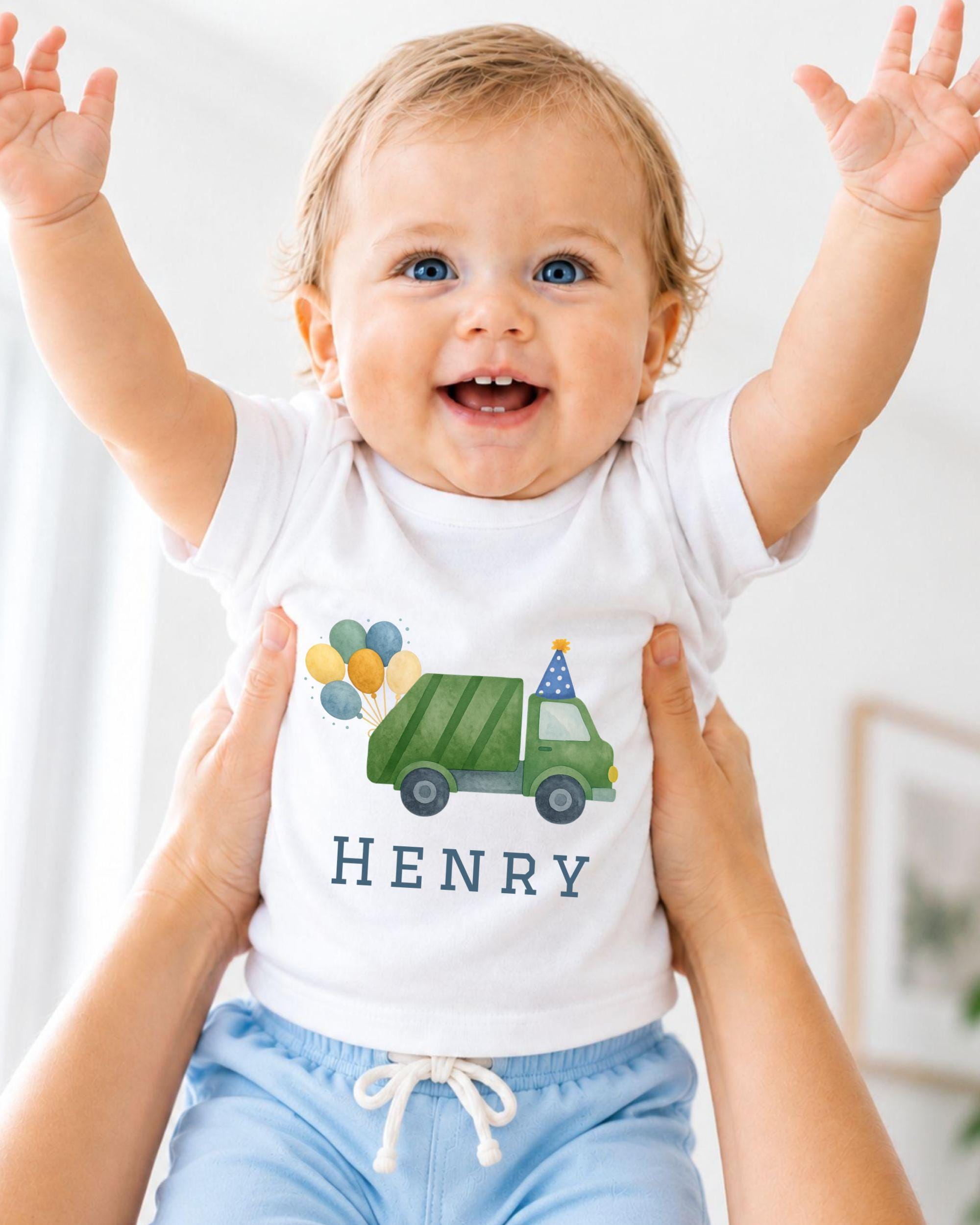 A smiling baby wearing a white shirt with a green truck and balloons on it, being held up by someone's arms.