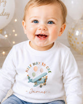 A smiling baby boy wearing a white t-shirt with a graphic of a plane and the text "Half way to one" and "James" printed on it.