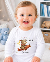 A smiling baby wearing a white onesie with a cartoon bear and airplane design, sitting in a room with bookshelves and a teddy bear.