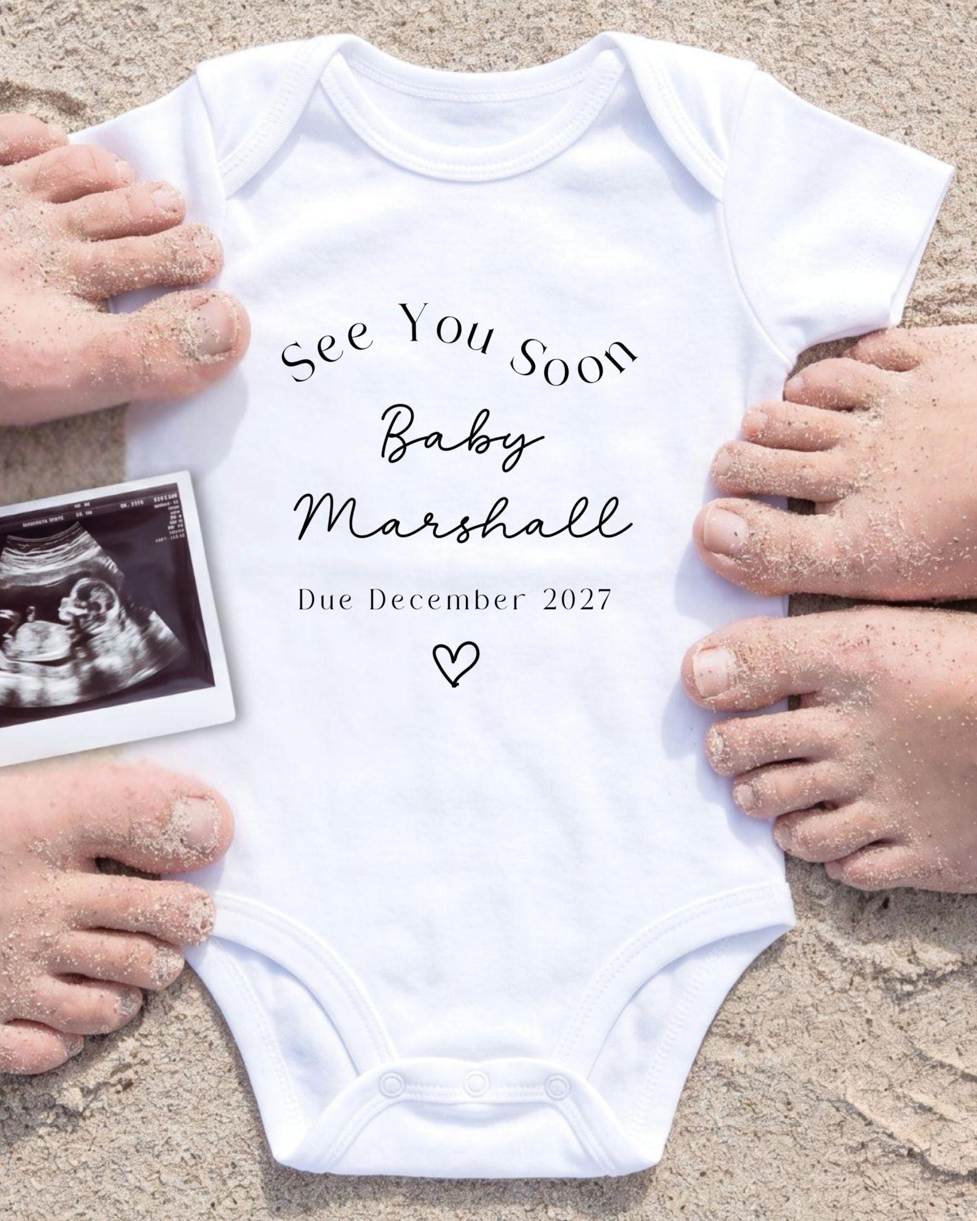 a white onesie with text and an image of an Ultrasound scan of a baby, surrounded by hands on a sandy beach.