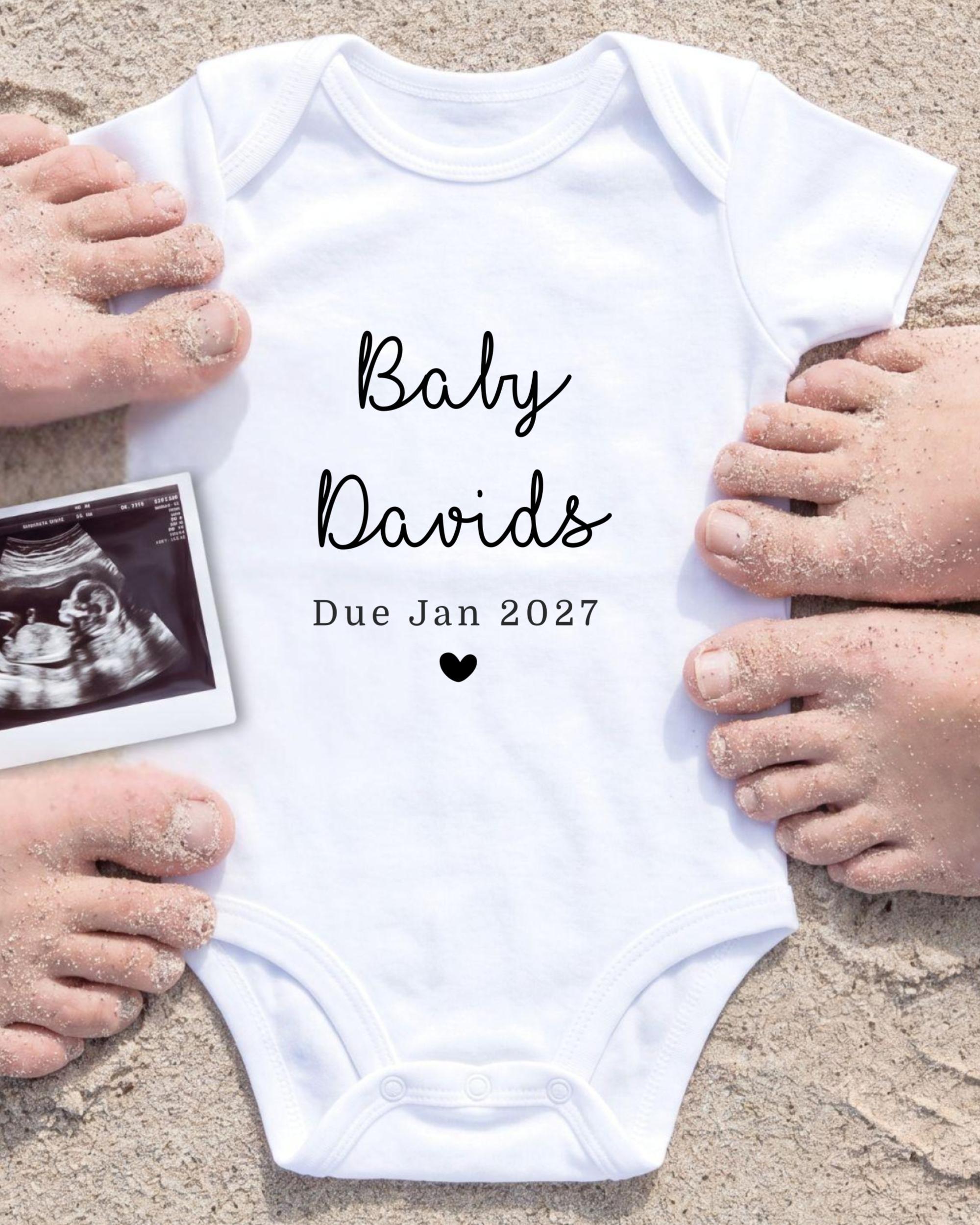 a white baby onesie with the text "Baby Davis Due Jan 2027" printed on it, surrounded by sand and a black and white photograph of a baby's belly.