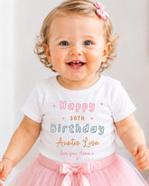 A smiling baby girl wearing a white t-shirt with text and a pink bow, standing in front of a white background.