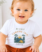A smiling baby wearing a white t-shirt with a camping-themed graphic and text.