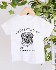 A white t-shirt with a graphic of a dog's head and the text "PROTECTED BY Cooper" printed on it, hanging on a wooden hanger.