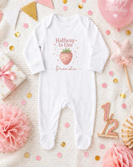 A white onesie with a strawberry design and the text "Halfway to One" printed on it, surrounded by pink and gold confetti, a pink star, and a pink balloon with the text "Happy Birthday" written on it.