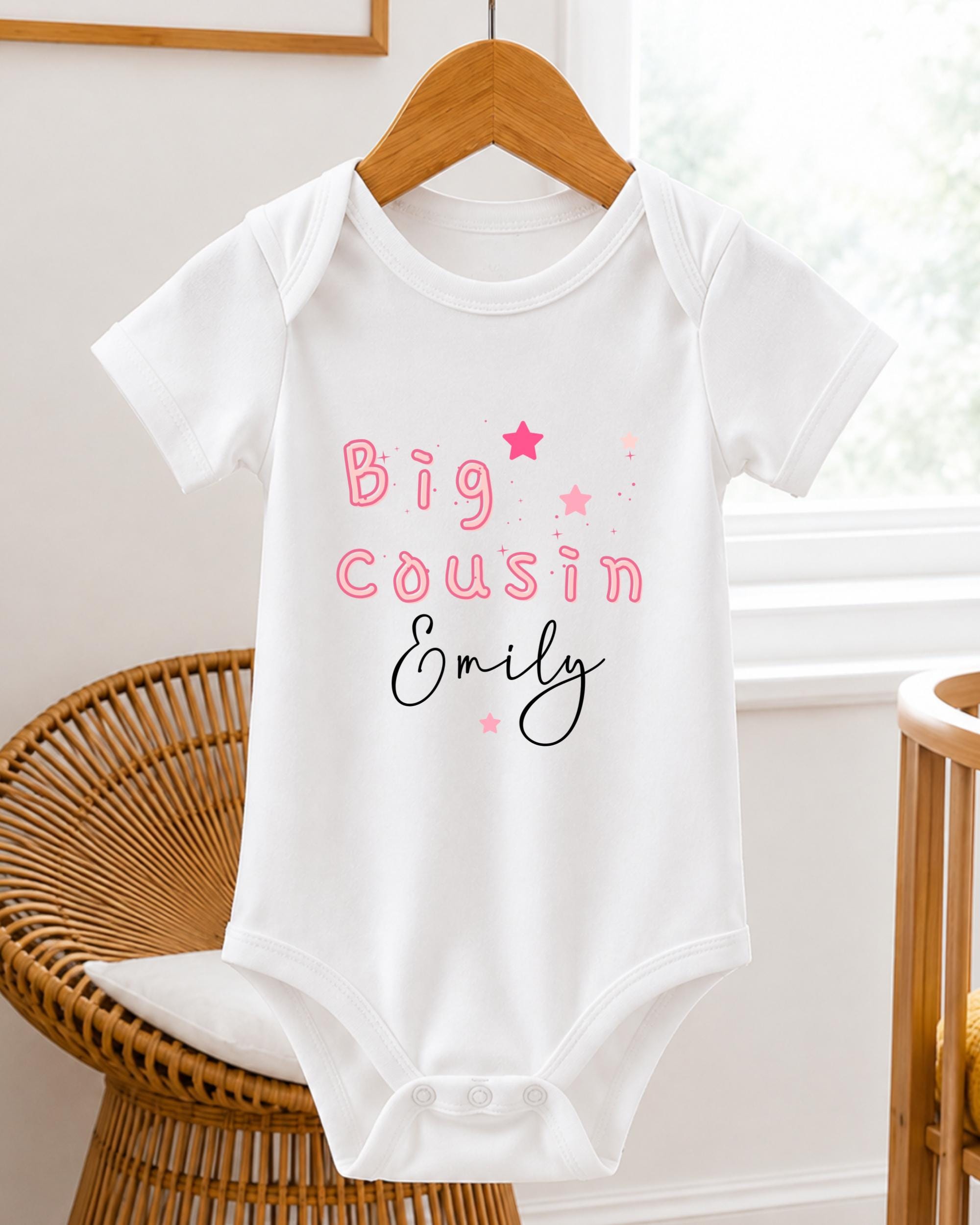 A white baby onesie with the text "Big Cousin Emily" printed on it, hanging on a wooden hanger.
