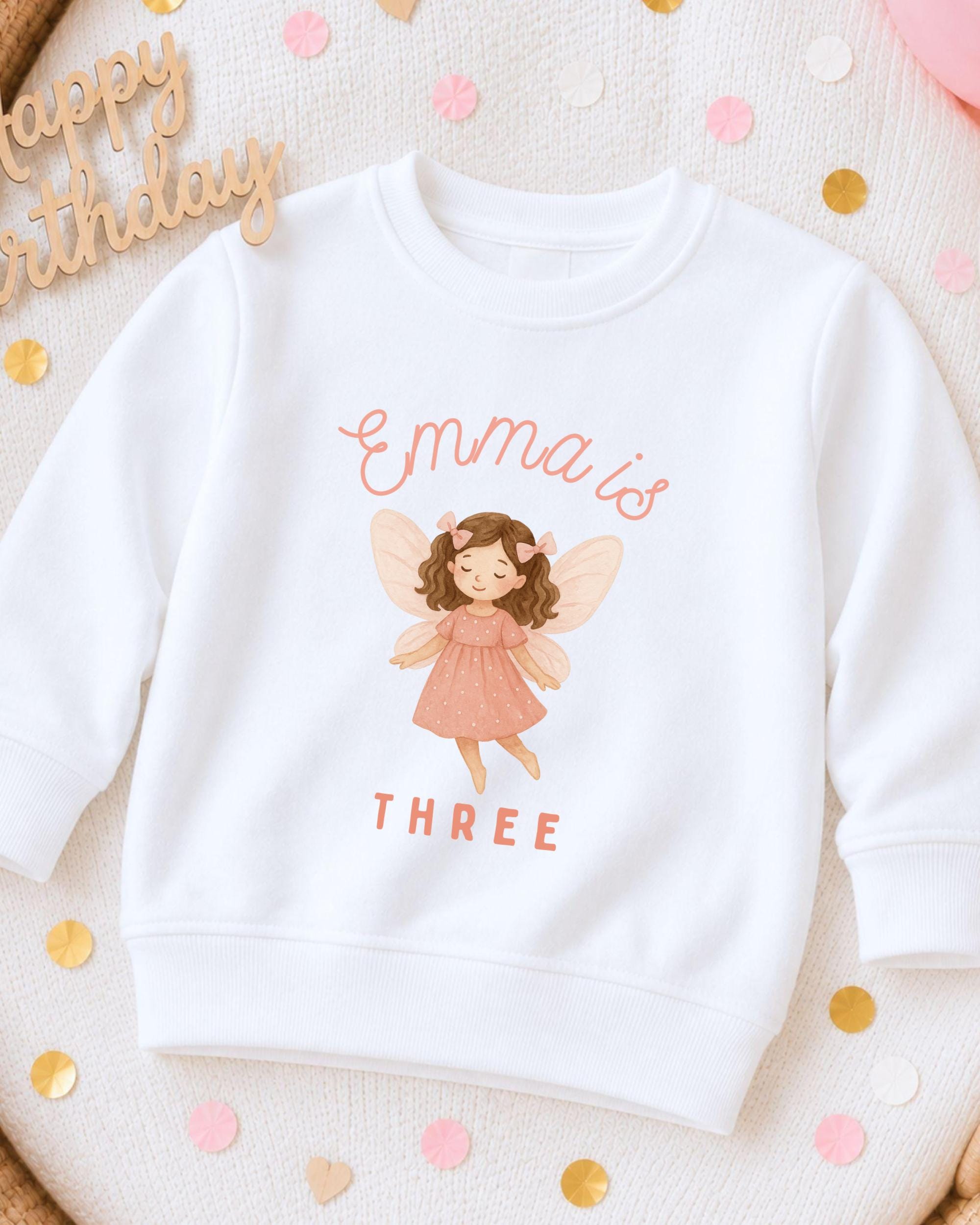 A white sweatshirt with a graphic of a fairy and the text "Emma is THREE" printed on it, surrounded by confetti.
