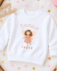 A white sweatshirt with a graphic of a fairy and the text "Emma is THREE" printed on it, surrounded by confetti.
