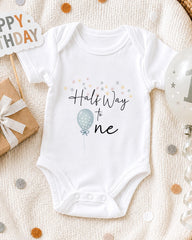 A white onesie with the text "Half Way to One" printed on it, along with a balloon and confetti, suggesting a celebration or milestone related to a baby's first birthday.