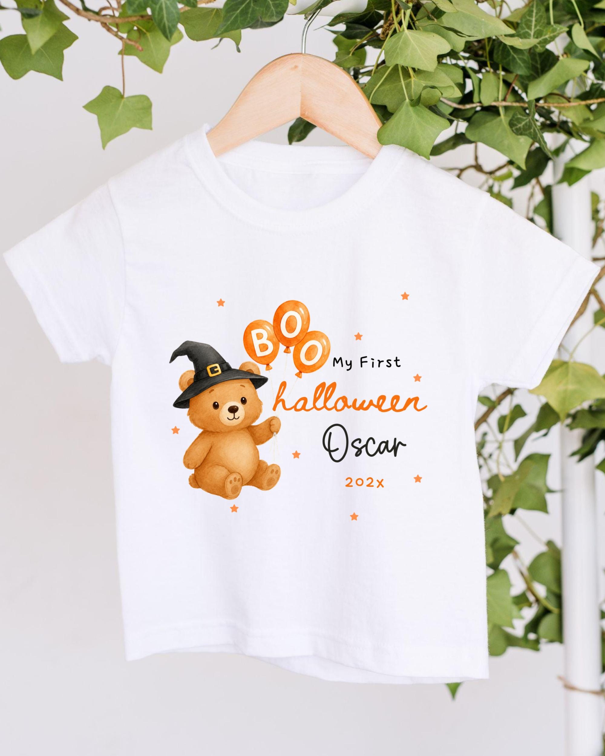A white t-shirt with a cartoon image of a teddy bear wearing a witch's hat and surrounded by orange balloons, along with text that reads "My First Halloween Oscar 202x".