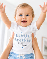 A smiling baby wearing a white shirt with the text "Little Brother Tho" is being held up by someone's arms.